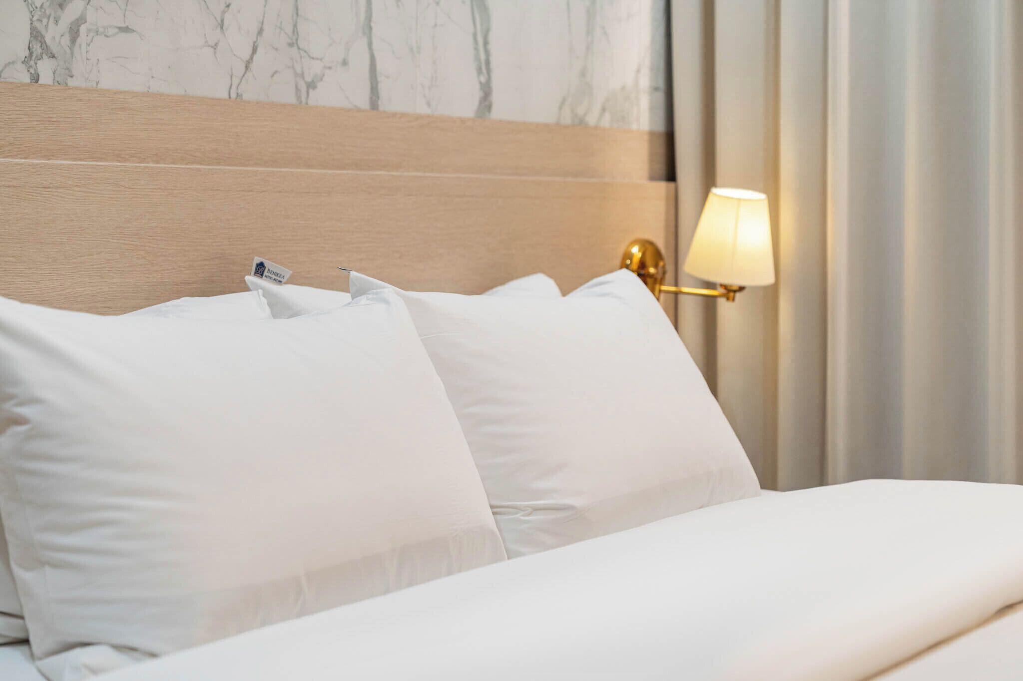 A neatly made bed with white linens and a wooden headboard. A gold lamp is mounted on the wall beside the bed.