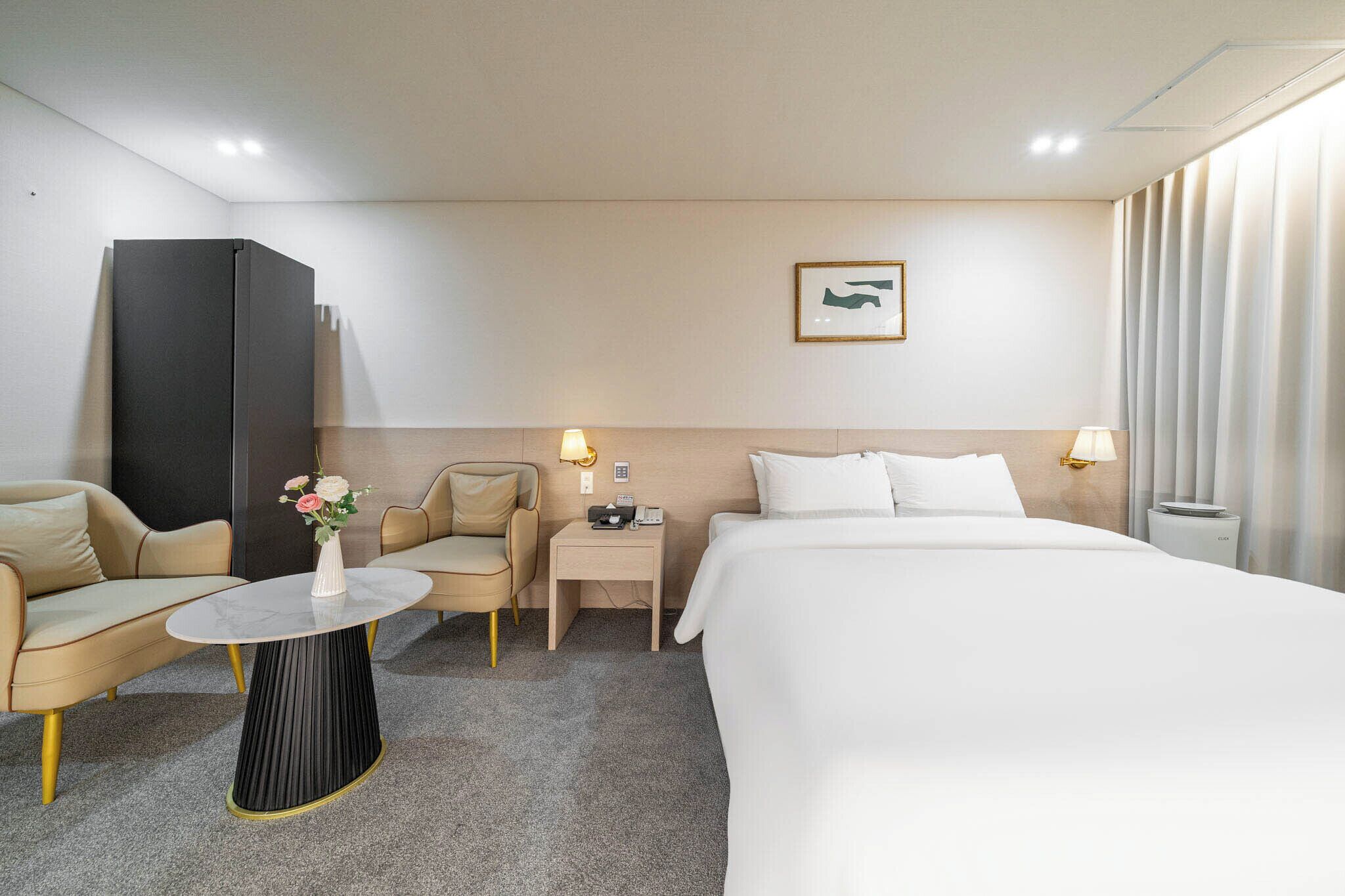 Habitación de hotel con cama, sofá, una mesita con un florero y una lámpara en la mesita de noche, además de un cuadro en la pared.