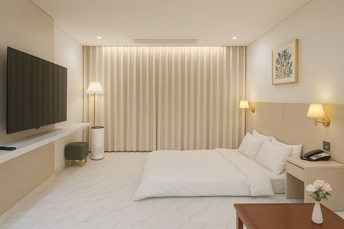 A modern hotel room with a flat-screen TV, a standing lamp, a bedside table with a phone, and a vase of flowers.