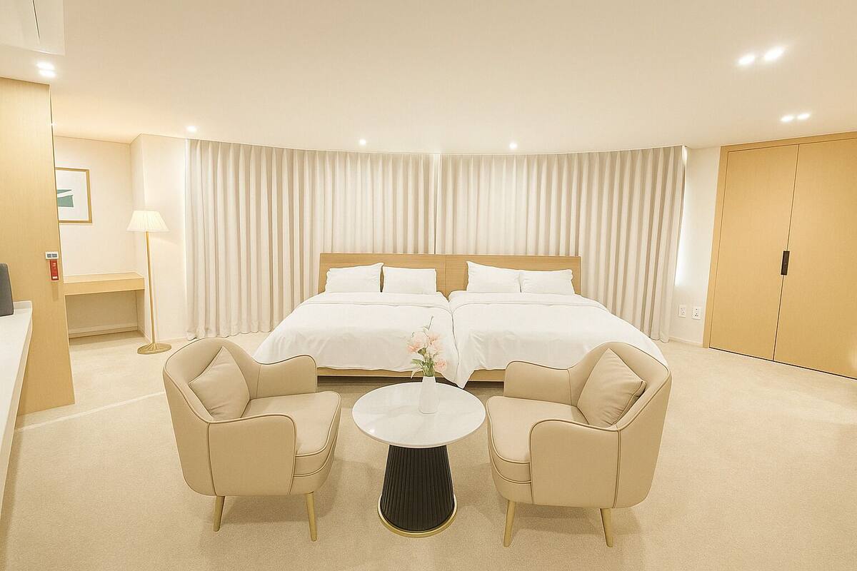 A hotel room with two beds, a round table, two armchairs, a desk, and a lamp.