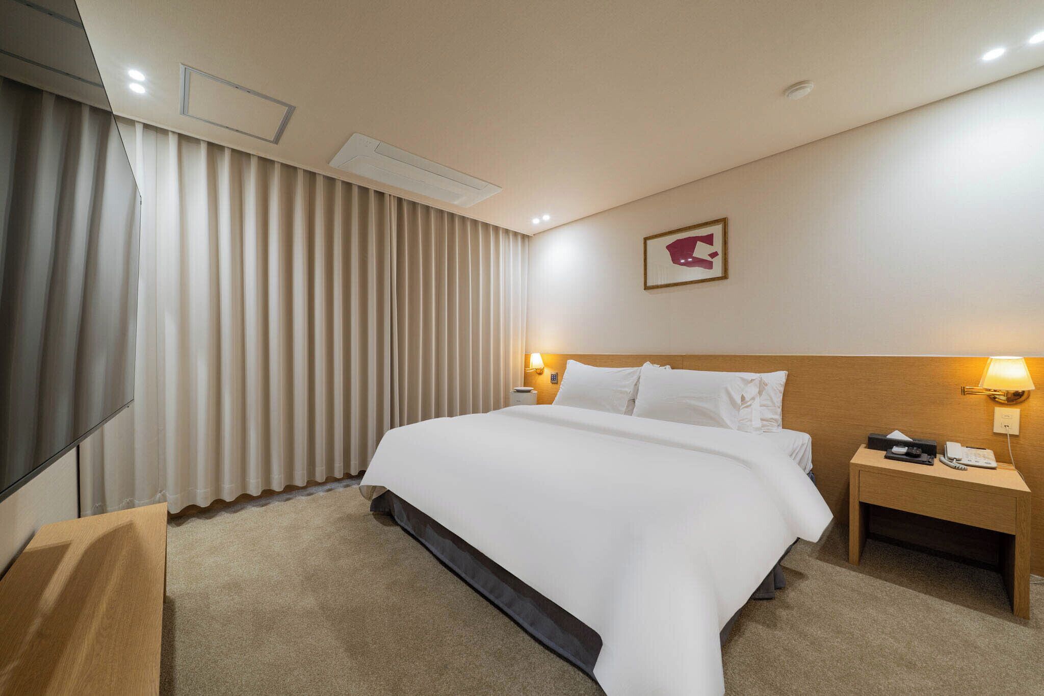 Habitación de hotel con una cama grande, una mesita de noche con lámpara, un televisor de pantalla plana y un amplio ventanal con cortinas.