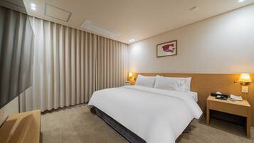 Habitación de hotel con una cama grande, una mesita de noche con lámpara, un televisor de pantalla plana y un amplio ventanal con cortinas.