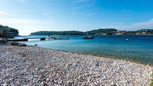 Une plage de galets bordée d’une eau claire, avec un quai et des collines qui se dessinent à l’horizon.