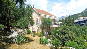 Una casa de dos pisos con techo de tejas rojas, rodeada de abundante vegetación y terreno pedregoso.