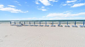 A beach with rows of blue beach chairs and umbrellas, a clear sky, and the ocean in the background.