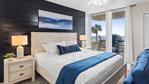 A bedroom with a bed, nightstands, a ceiling fan, a painting, and a balcony with a view.