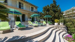 Ein sonniger Außenbereich mit grünen Lounge-Sesseln mit Muster, Sonnenschirmen, einer Steintreppe und Bergblick.