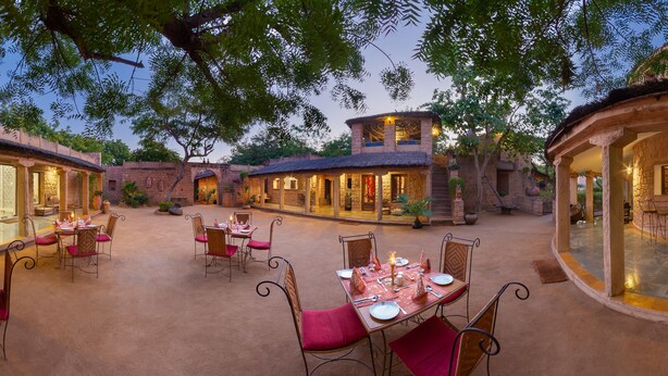 An outdoor dining area with stone walls and tables set for dining, surrounded by trees and a stone building in the background. - MANVĀR SHERGARH, THE DESERT RESORT (Shergarh)