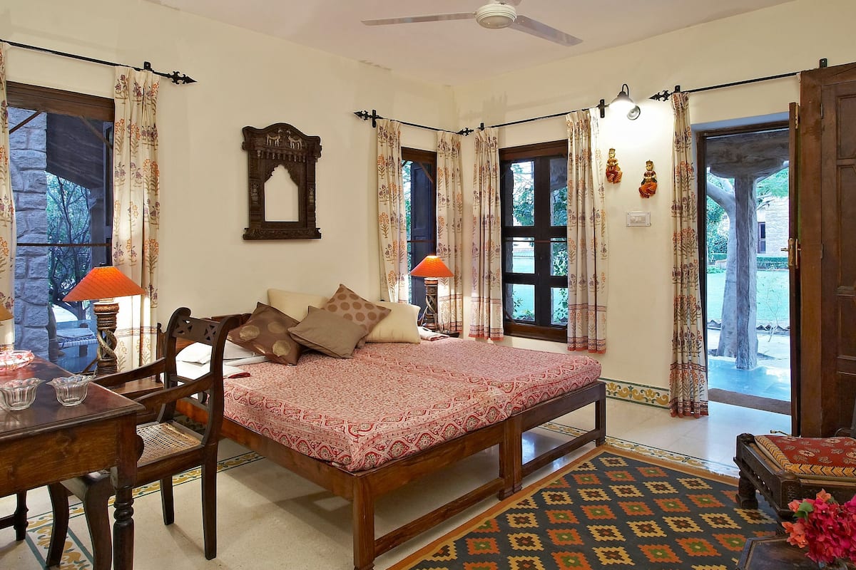 Quarto tradicional com cama, escrivaninha e cadeira. Cortinas, espelho e porta que dá para o exterior.
