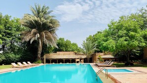 A pool area with lounge chairs, a palm tree, and a building with a thatched roof. - MANVĀR SHERGARH, THE DESERT RESORT (Shergarh)