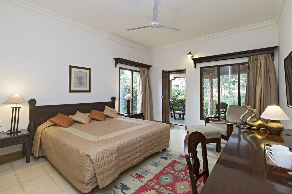 Quarto de hotel com uma cama grande, duas mesas de cabeceira com abajures, uma escrivaninha com cadeira e vista para o exterior através de uma porta de vidro.