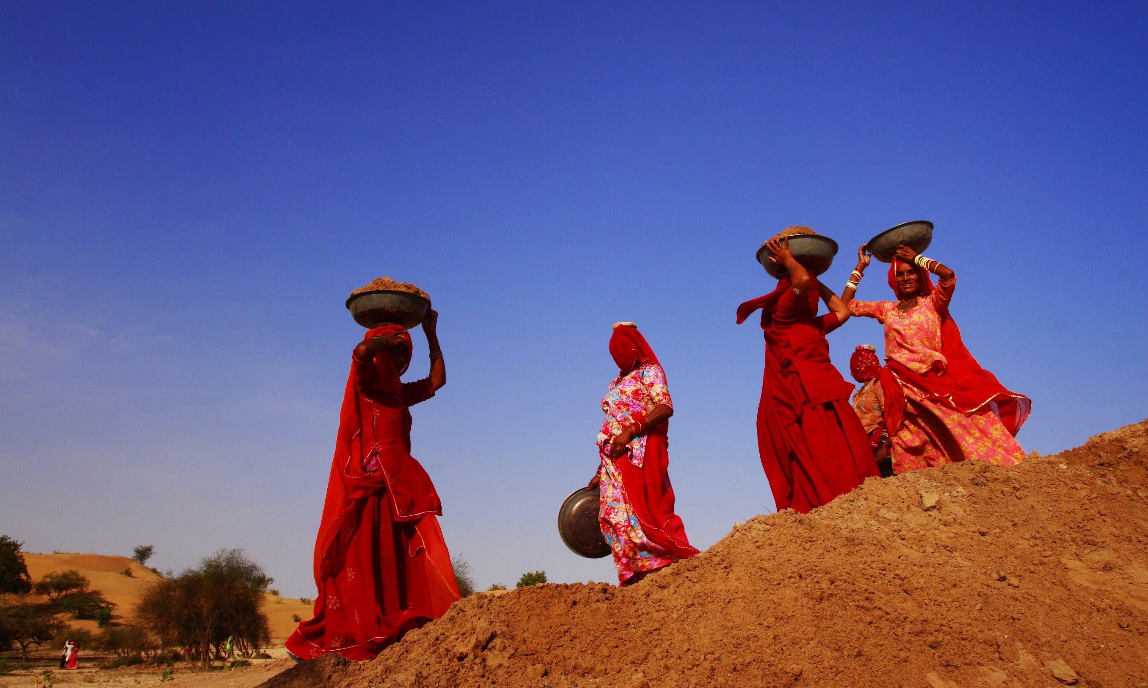 A group of people in traditional attire carrying large containers on their heads, walking on a sandy path.