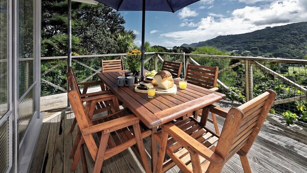 Una terraza de madera con una mesa, sillas, un paraguas y una vista panorámica de montañas y vegetación.