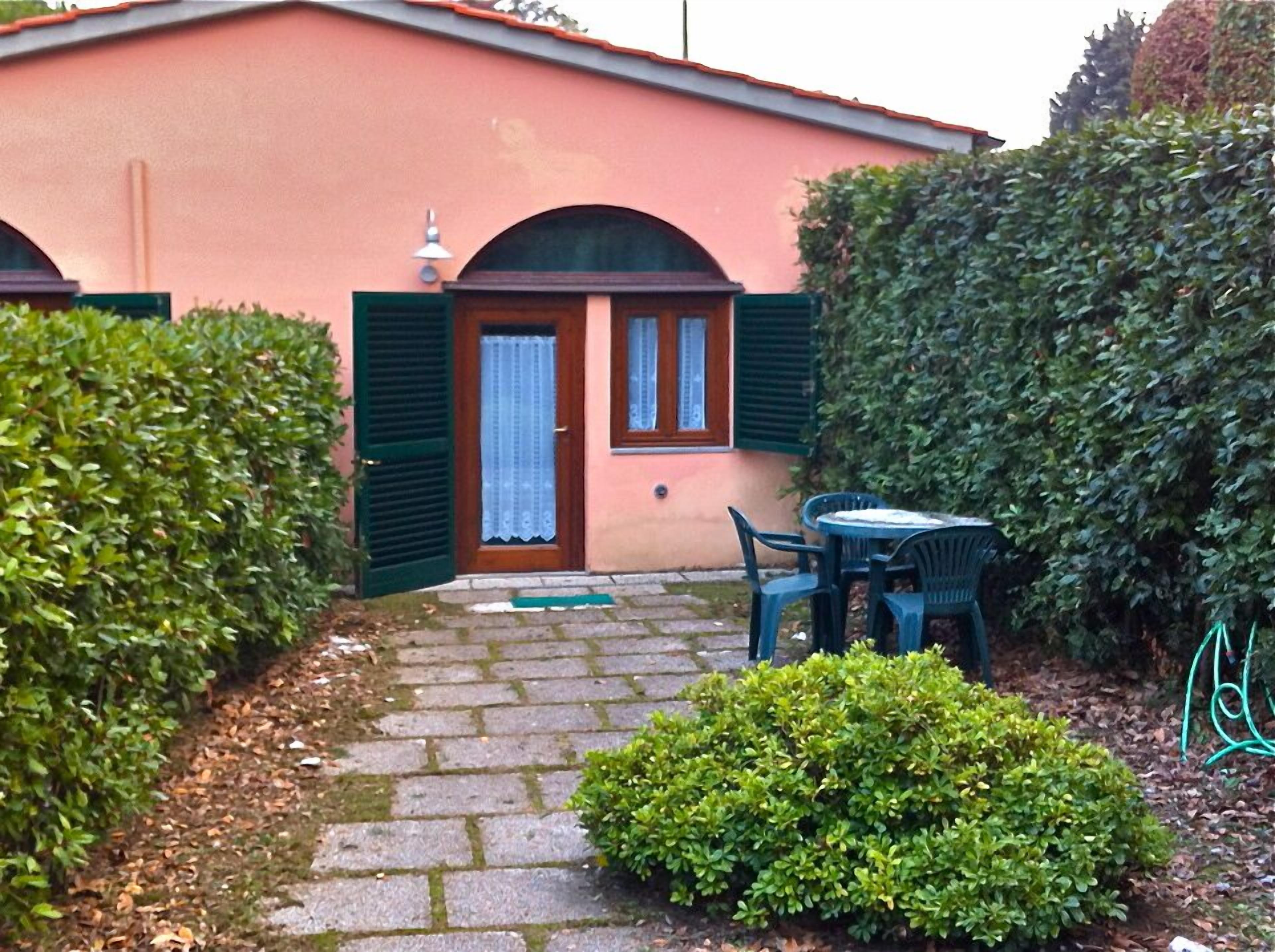 Un pequeño edificio con un sendero pavimentado que lleva a una puerta con contraventanas verdes. Hay una mesa y sillas afuera.