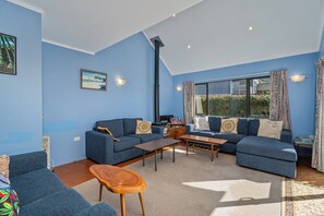 A living room with blue walls, a sofa set, a wooden coffee table, and a fireplace. - Rosemary Cottage - Hahei Holiday Home (Hahei)