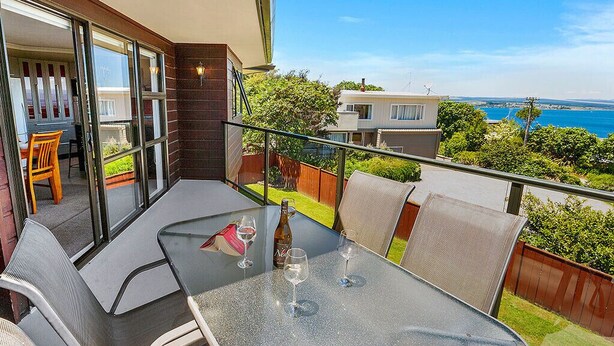 A balcony with a glass table and chairs, a bottle of wine, and a view of the sea. - Acacia Bay Getaway - Lake Taupo Holiday Home (Acacia Bay)