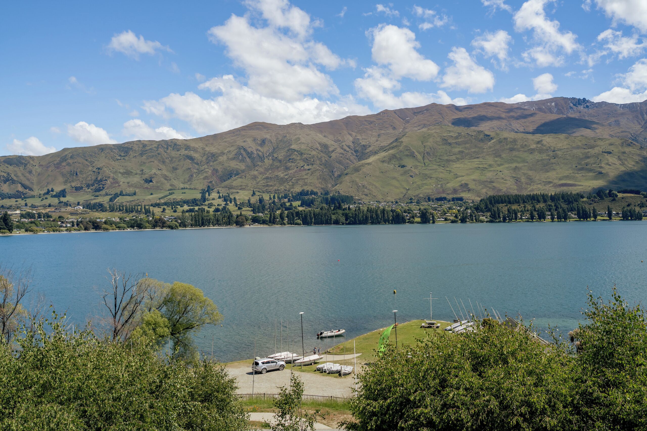 A lake with mountains in the background, a parking area with cars, and a clear sky.