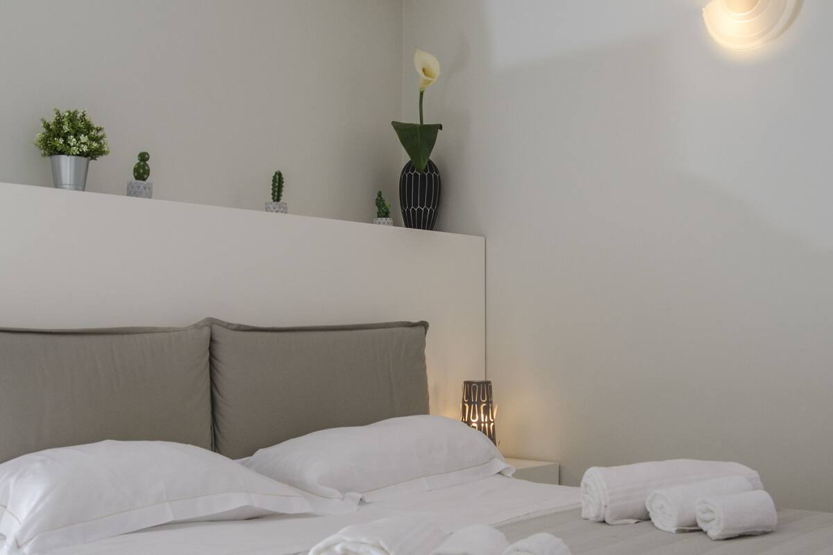 A modern hotel room with a large bed, a bedside table, a wall-mounted light, and a shelf with decorative items.