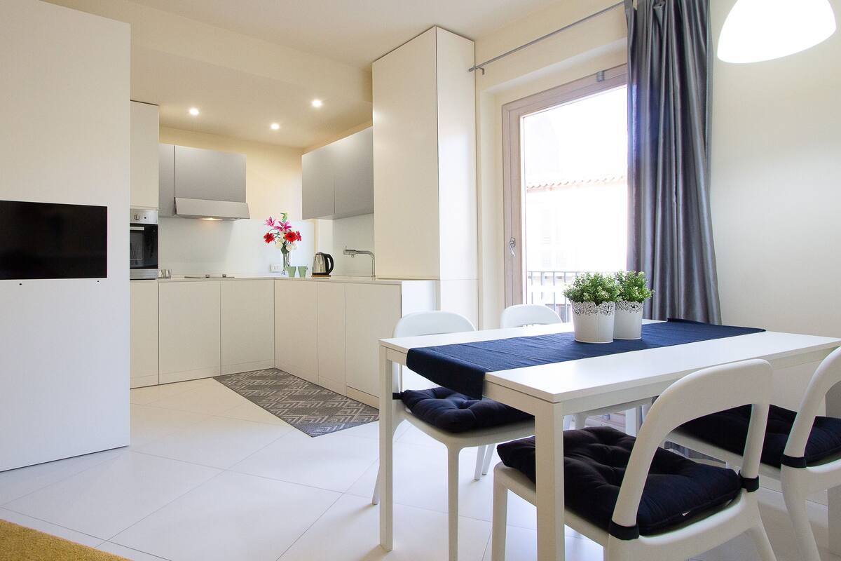 A modern kitchen with white cabinets, a built-in oven, and a coffee maker. There is a dining table with two chairs and a blue tablecloth.