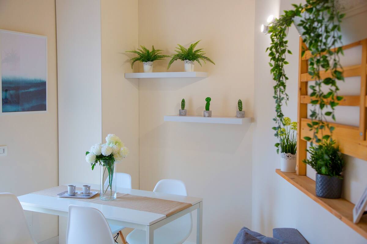 A modern dining area with white furniture, a vase with white flowers, and potted plants on shelves.