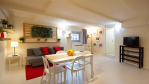 A modern living room with a white dining table, white chairs, a grey sofa with red cushions, a red rug, a TV on a black stand, and a green plant wall. - Stesicoro Exclusive House (Catania)