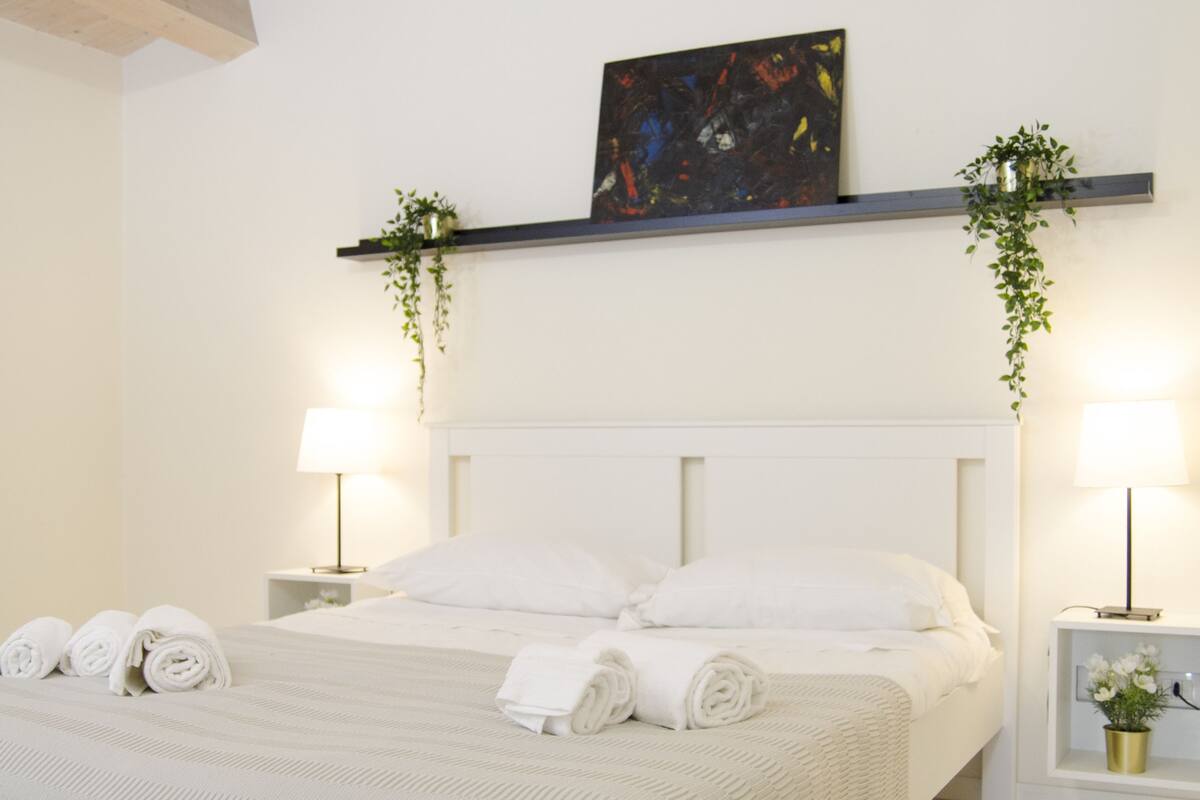 A neatly made bed with white linens, two bedside tables with lamps, and a painting above the bed.