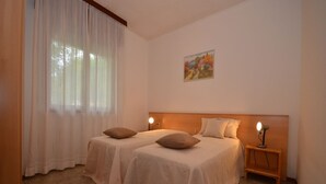 A room with two beds, a nightstand with a lamp, a painting on the wall, and a window with curtains. - Ville Annamaria (Lignano Sabbiadoro)