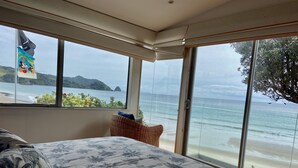 A bedroom with a large bed, a wicker chair, and a view of the beach and ocean through large windows. - Bliss on the Beach - Whangapoua (Whangapoua)