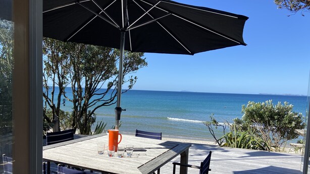 A wooden deck with a table and chairs, an umbrella, and a view of the ocean. - Bliss on the Beach - Whangapoua (Whangapoua)