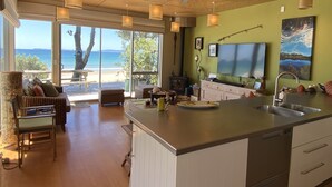 A modern kitchen with a large island, a dining area with a view of the beach, and a ceiling fan. - Bliss on the Beach - Whangapoua (Whangapoua)