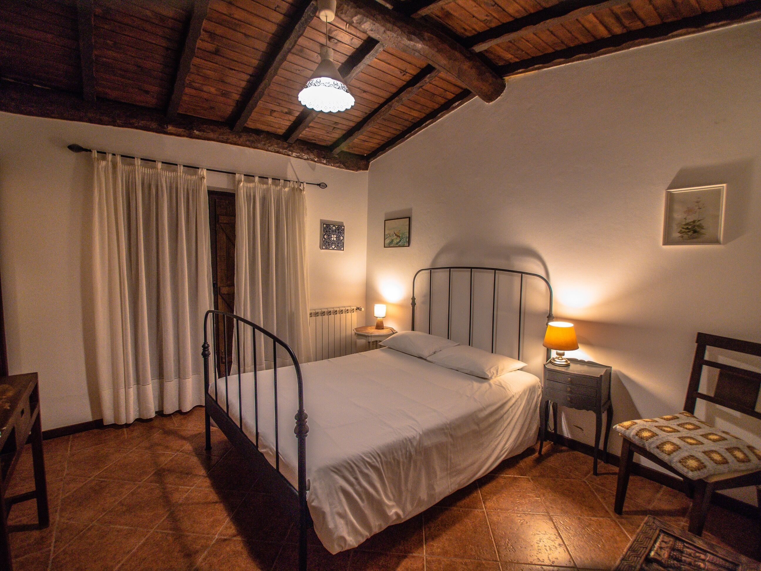 Ein Schlafzimmer mit einem Bett, einem Nachttisch mit Lampe, einem Sessel, einem Fenster mit Vorhängen und einer Holzbalkendecke.
