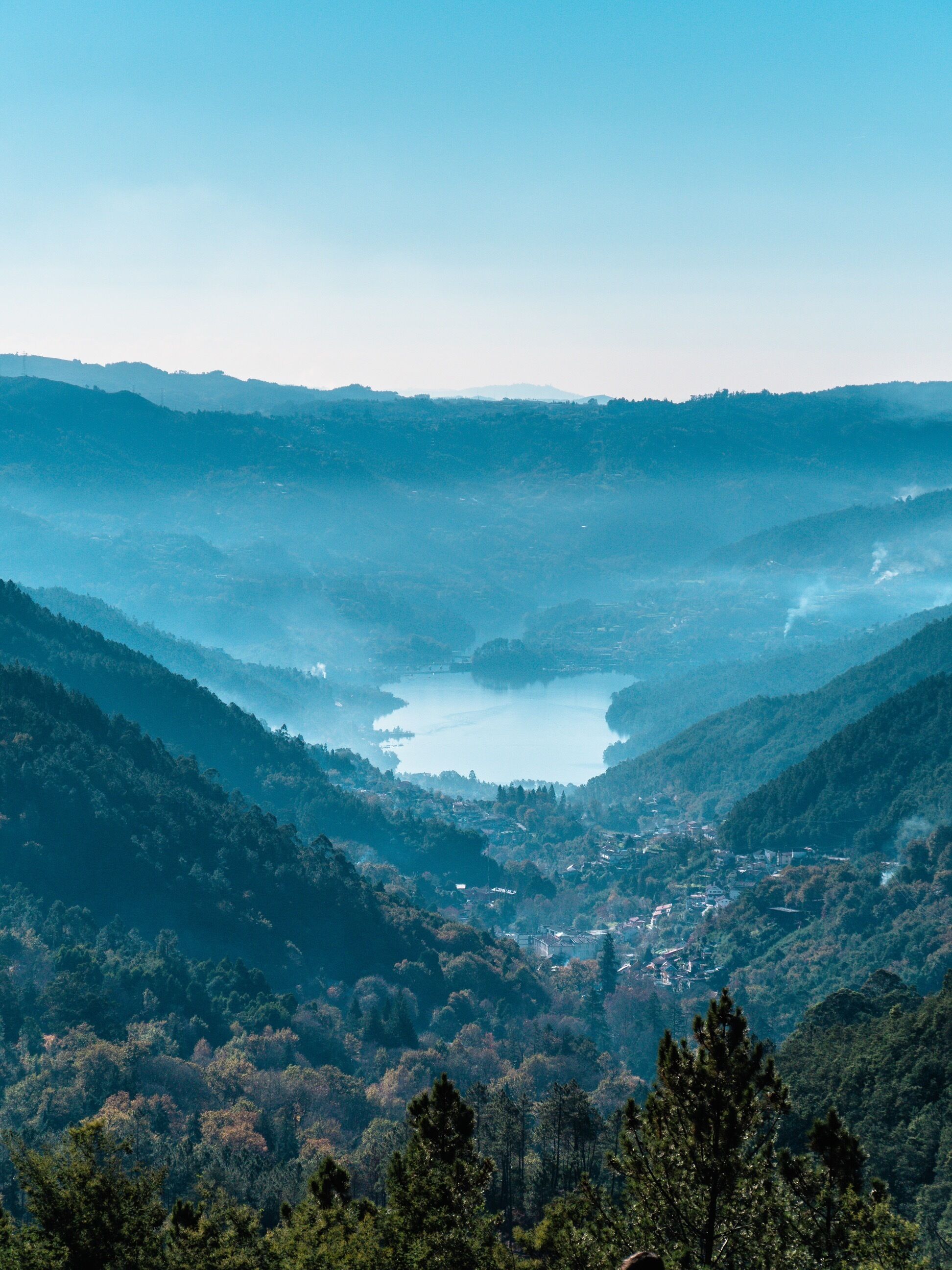A panoramic view of a valley with a lake, surrounded by forested hills.