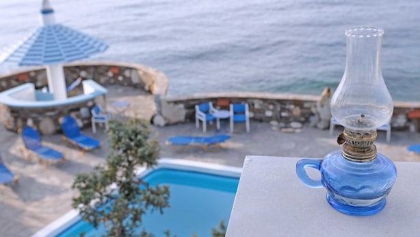 A poolside view with a clear view of the ocean, a lounge area with blue chairs, and a decorative glass lamp. - Daidalos Hotel (Ikaria)
