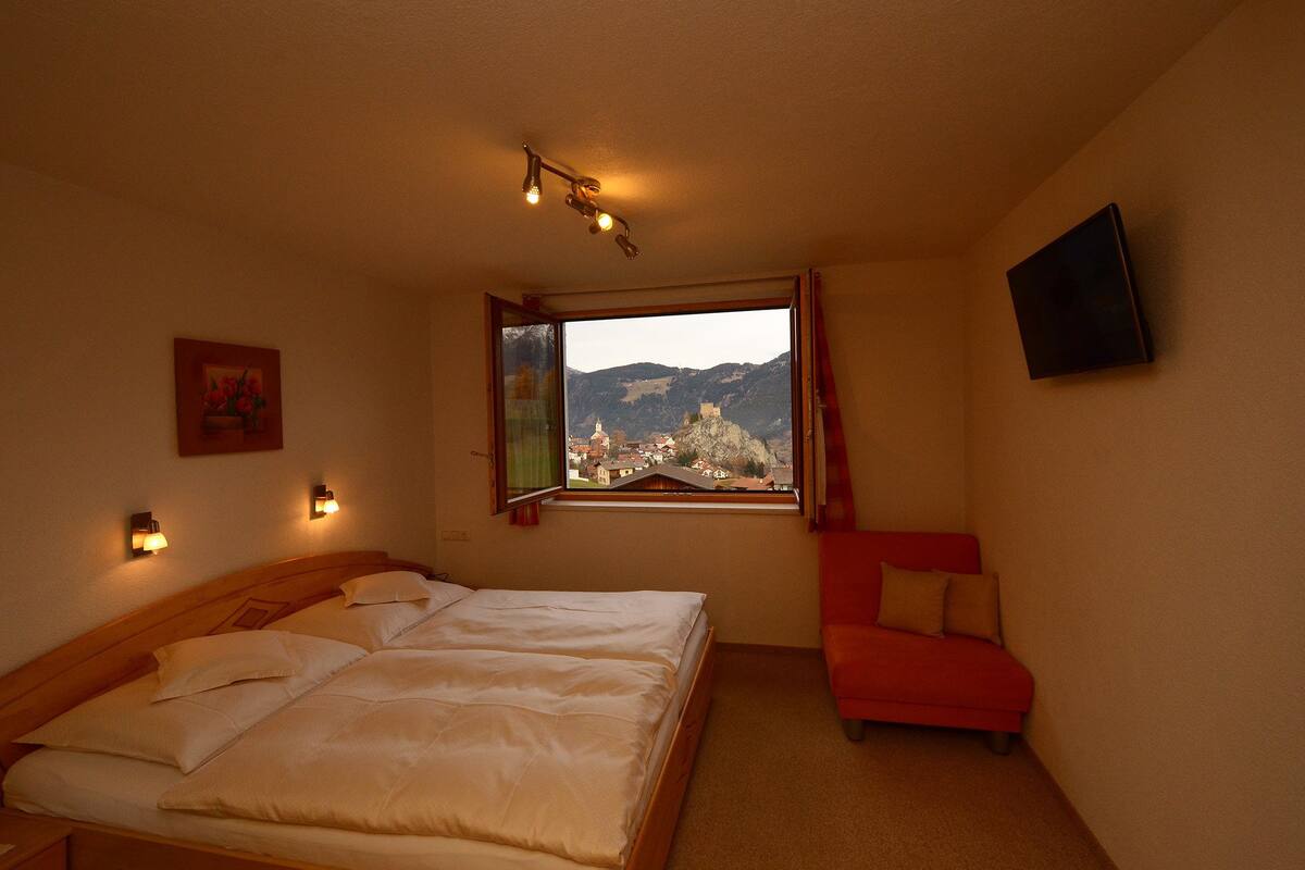 Un dormitorio con una cama, una silla roja, un ventanal con vistas a la montaña y luces fijadas a la pared.