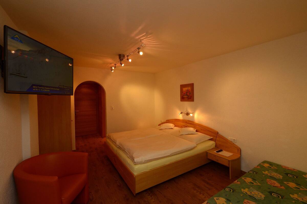 Un dormitorio con cama, televisor montado en la pared, mesita de noche, silla y una pequeña mesa con lámpara.