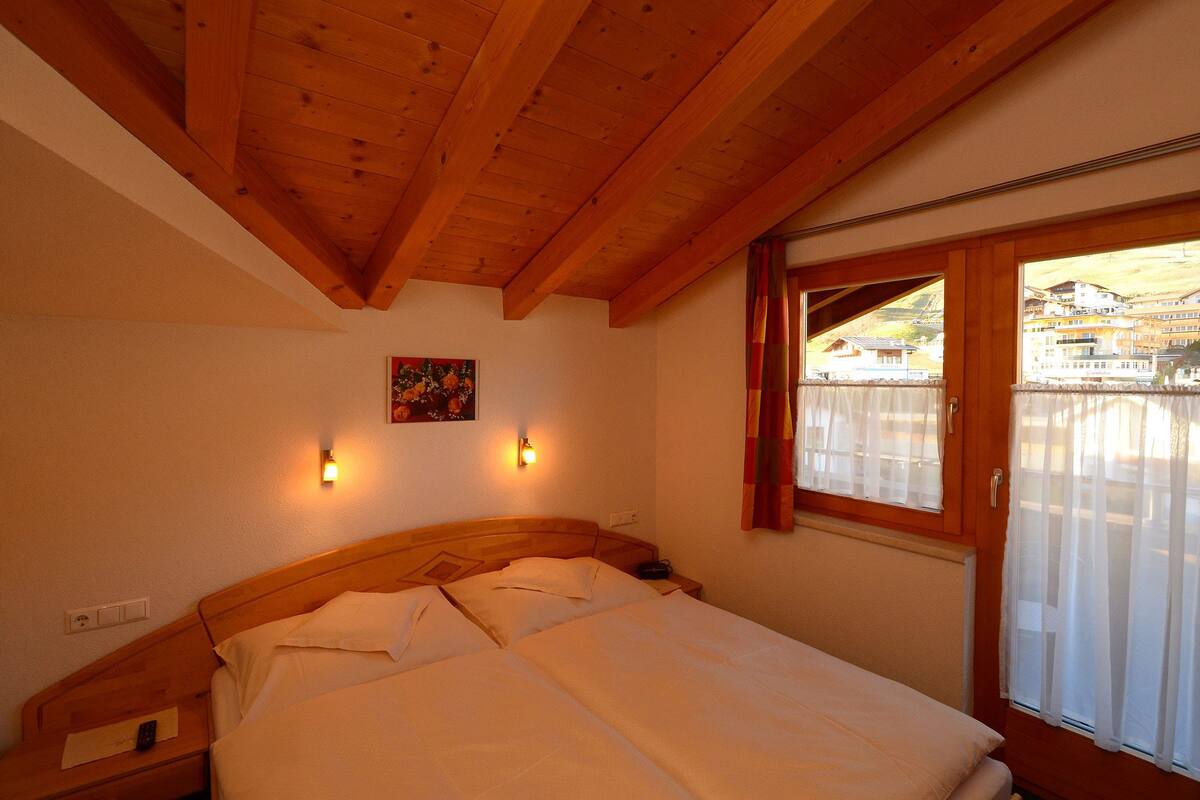 Un dormitorio con techo de madera, cama con sábanas blancas, dos lámparas de pared y una ventana con cortinas.