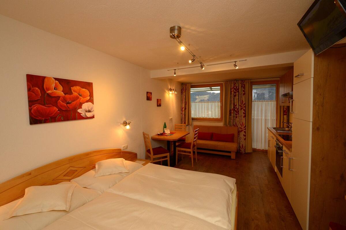 Habitación de hotel con cama, mesa de comedor con sillas, una pequeña cocina y televisor montado en la pared.