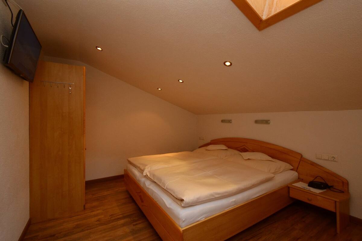 Un dormitorio con una cama de madera, una mesita de noche, un armario de madera y un televisor montado en la pared.