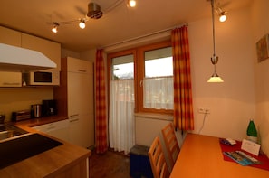 A compact kitchen with a microwave, stove, and wooden cabinets. There is a dining table with chairs and a window with curtains. - Edelweiss Ladis (Ladis)