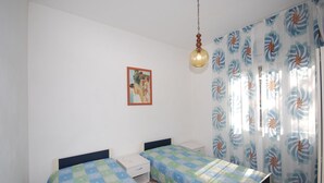 A bedroom with two beds, a nightstand, a hanging light fixture, a framed picture on the wall, and a window with floral curtains. - Ville Canarino (Lignano Sabbiadoro)