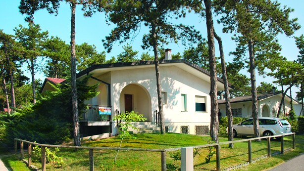 A small, single-story house with a red roof, surrounded by trees and a fenced yard. - Ville Canarino (Lignano Sabbiadoro)