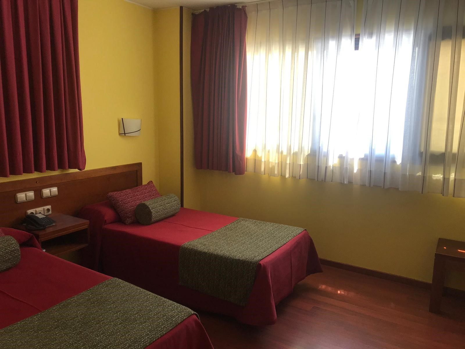 Een hotelkamer met twee bedden, een houten hoofdbord, een nachtkastje en een raam met gordijnen.