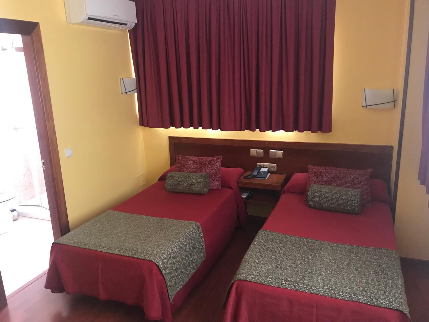 A hotel room with two single beds, each with a red bedspread and a grey blanket. There is a wooden nightstand with a telephone and a small table lamp. The room has a yellow wall and a window with red curtains.