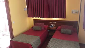 A hotel room with two single beds, each with a red bedspread and a grey blanket. There is a wooden nightstand with a telephone and a small table lamp. The room has a yellow wall and a window with red curtains.