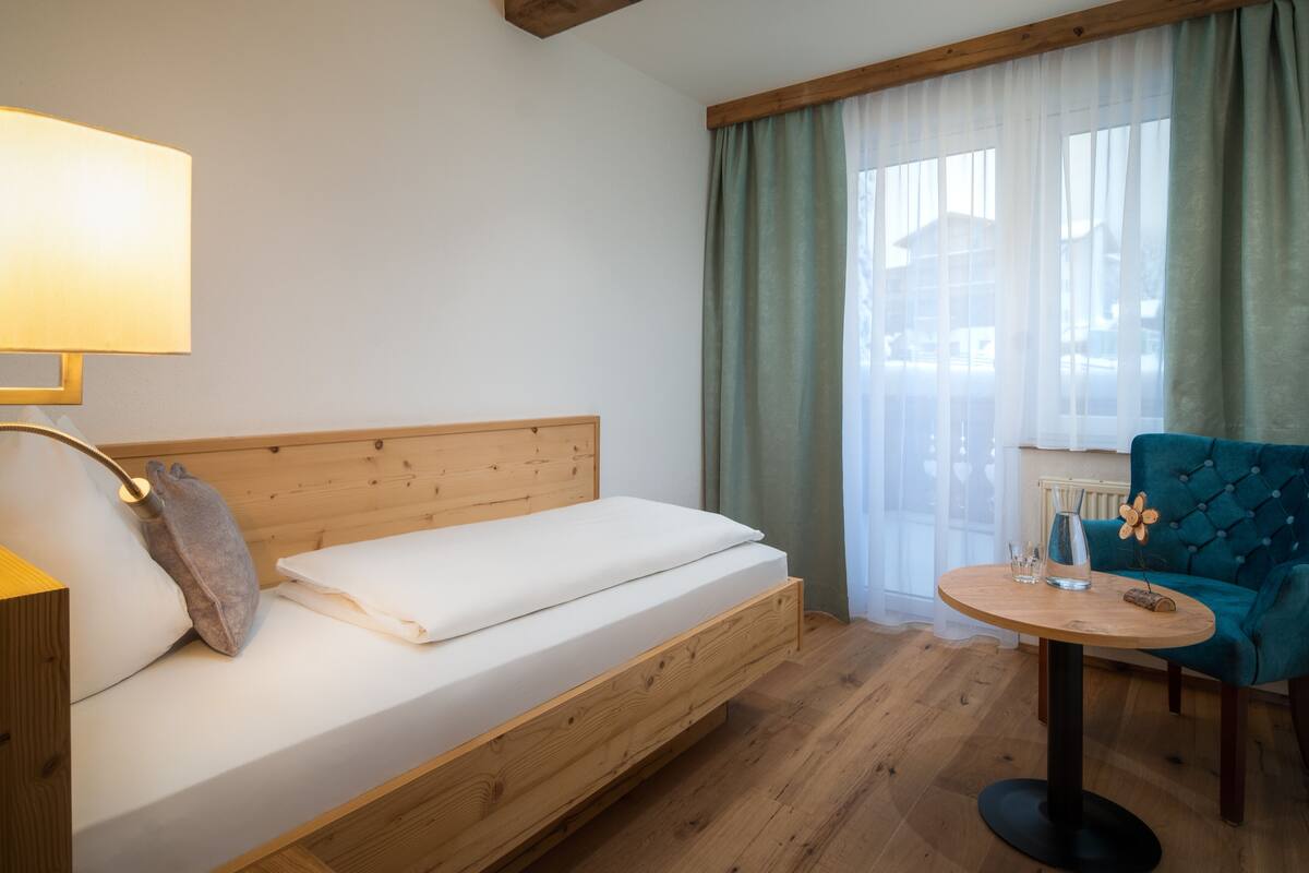 Ein Zimmer mit einem hölzernen Bett, einem runden Tisch mit Stuhl und einem Fenster mit Vorhängen.