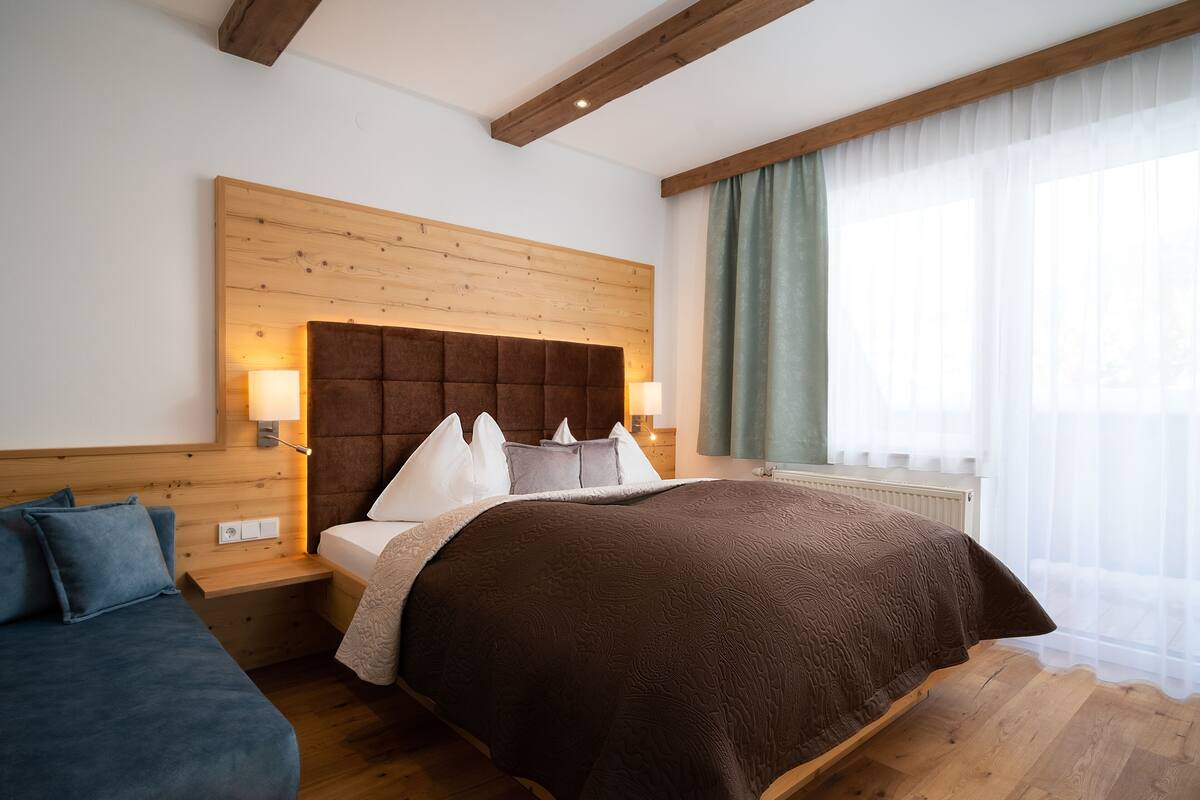Ein Hotelzimmer mit einem großen Bett, einem hölzernen Kopfteil, zwei Nachttischen mit Lampen, einem Beistelltisch und einem Fenster mit durchscheinenden Vorhängen.