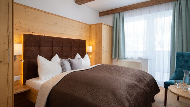 Ein Schlafzimmer mit einem großen Bett, einem hölzernen Kopfteil, Nachttischlampen, einem kleinen Tisch mit Gläsern und einem Fenster mit Vorhängen.
