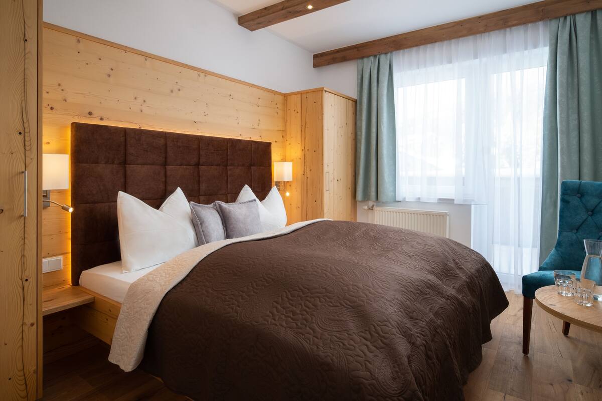 Ein Schlafzimmer mit einem großen Bett, einem hölzernen Kopfteil, Nachttischlampen, einem kleinen Tisch mit Gläsern und einem Fenster mit Vorhängen.