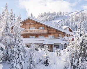 Eine Holzhütte namens BERGNEST mit einer Bar und einem Restaurant namens ZRYD, umgeben von schneebedeckten Bäumen und einer Skipiste im Hintergrund.