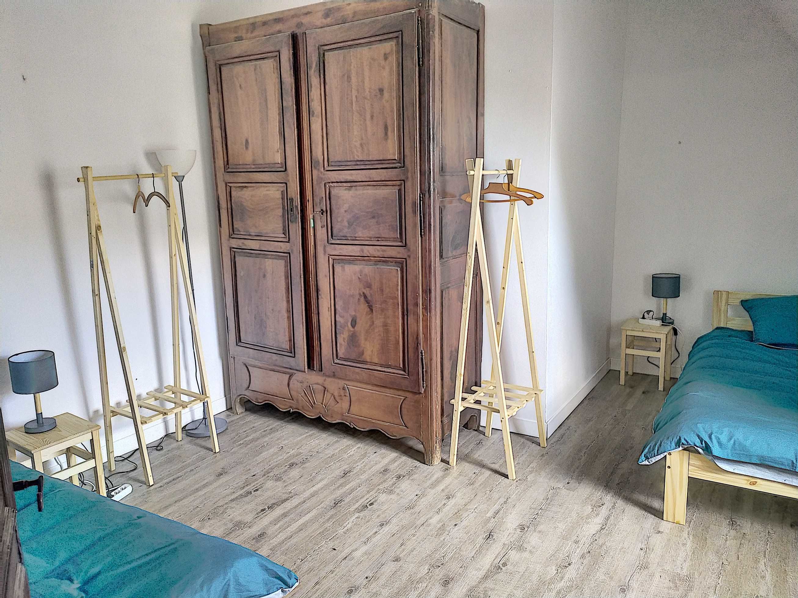 A room with a large wooden wardrobe, two wooden chairs, a small bedside table with a lamp, and a bed with a teal bedspread.